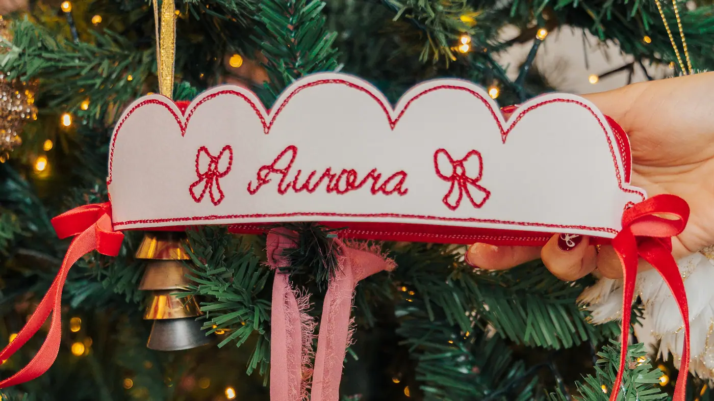 A scalloped white banner embroidered with a name, hanging on a lit Christmas tree with red ribbon