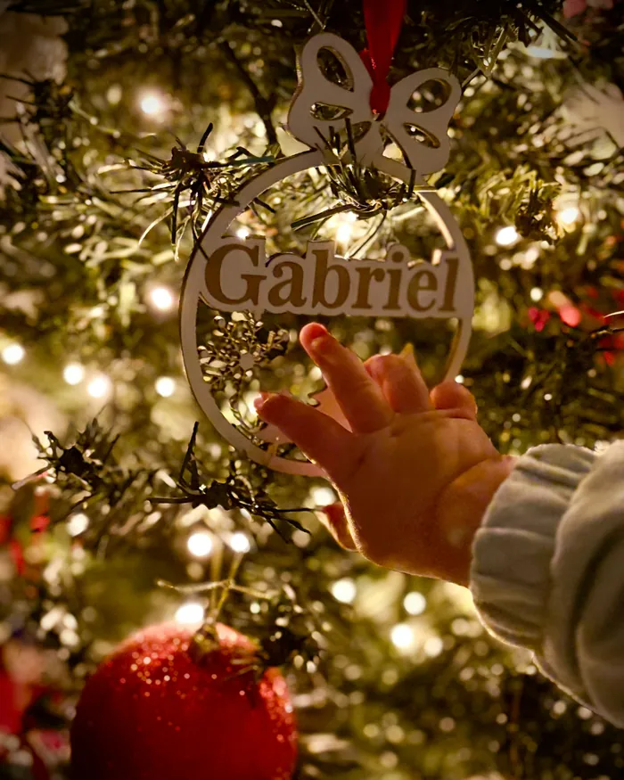 A baby's hand reaching for a gold ornament cut with the name Gabriel on a lit Christmas tree