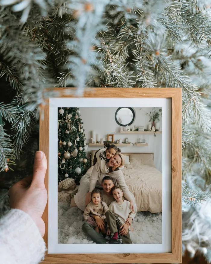 A hand holding a wooden frame against a frosted Christmas tree; inside, a photograph of parents and three children by a tree