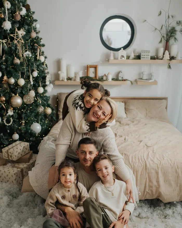 A mother and father sitting on the floor with their three children in front of a Christmas tree, all of them laughing