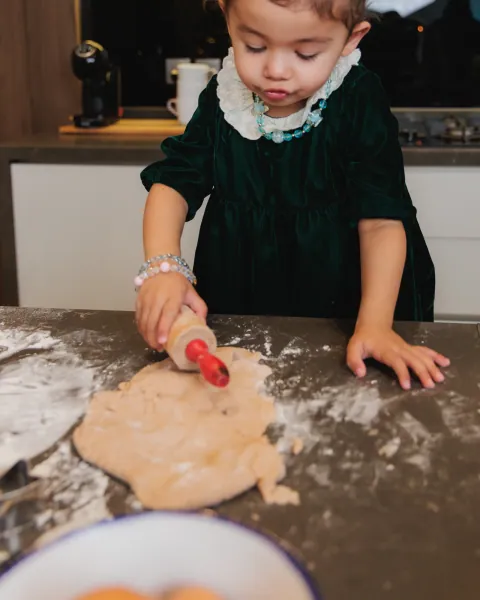 A small girl in a green velvet dress rolling gingerbread dough with a red rolling pin