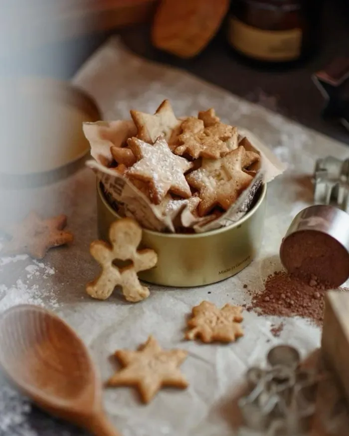 A small tin of star and gingerbread biscuits with a wooden spoon and cutters on a floured table