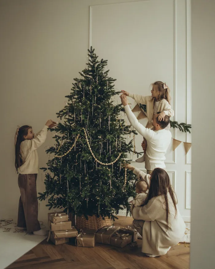 A mother and two girls decorating a tall real Christmas tree in a bright room with white walls