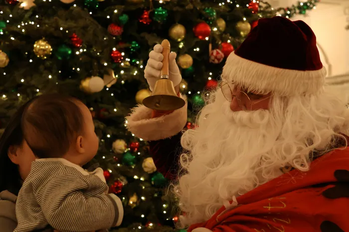Santa Claus ringing a small brass bell for a baby beside a decorated Christmas tree