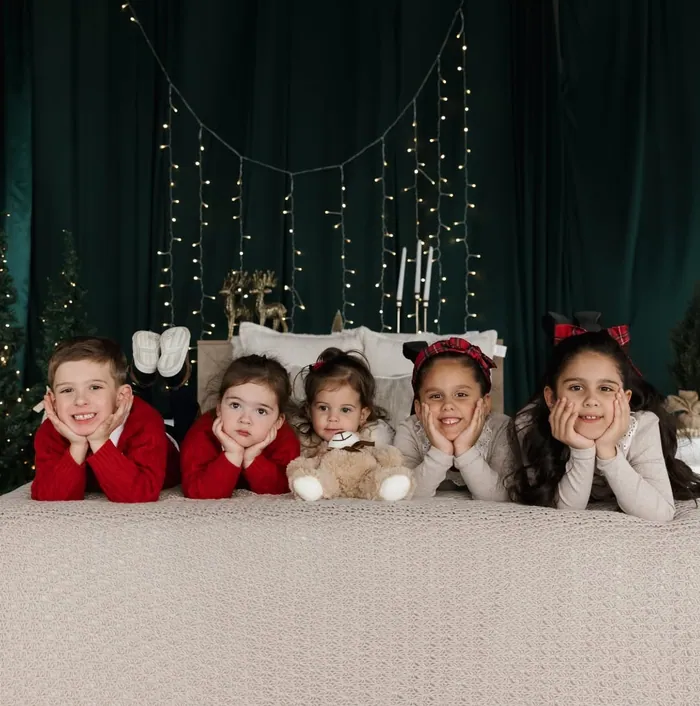 Five small children in red and cream jumpers lying in a row with their chins in their hands, fairy lights behind them