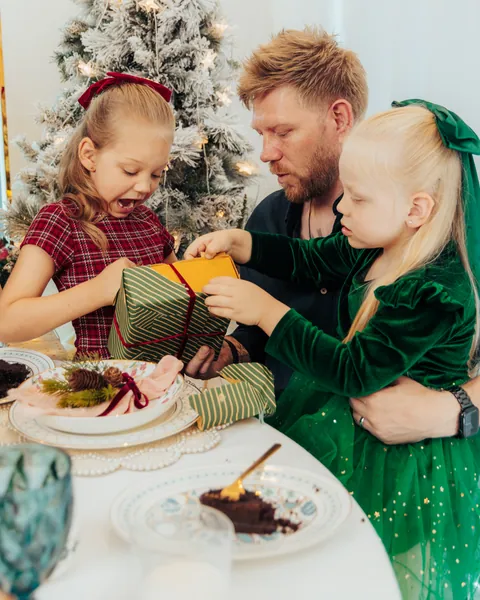 Two sisters in red tartan and green velvet opening a present with their father at a laid Christmas table