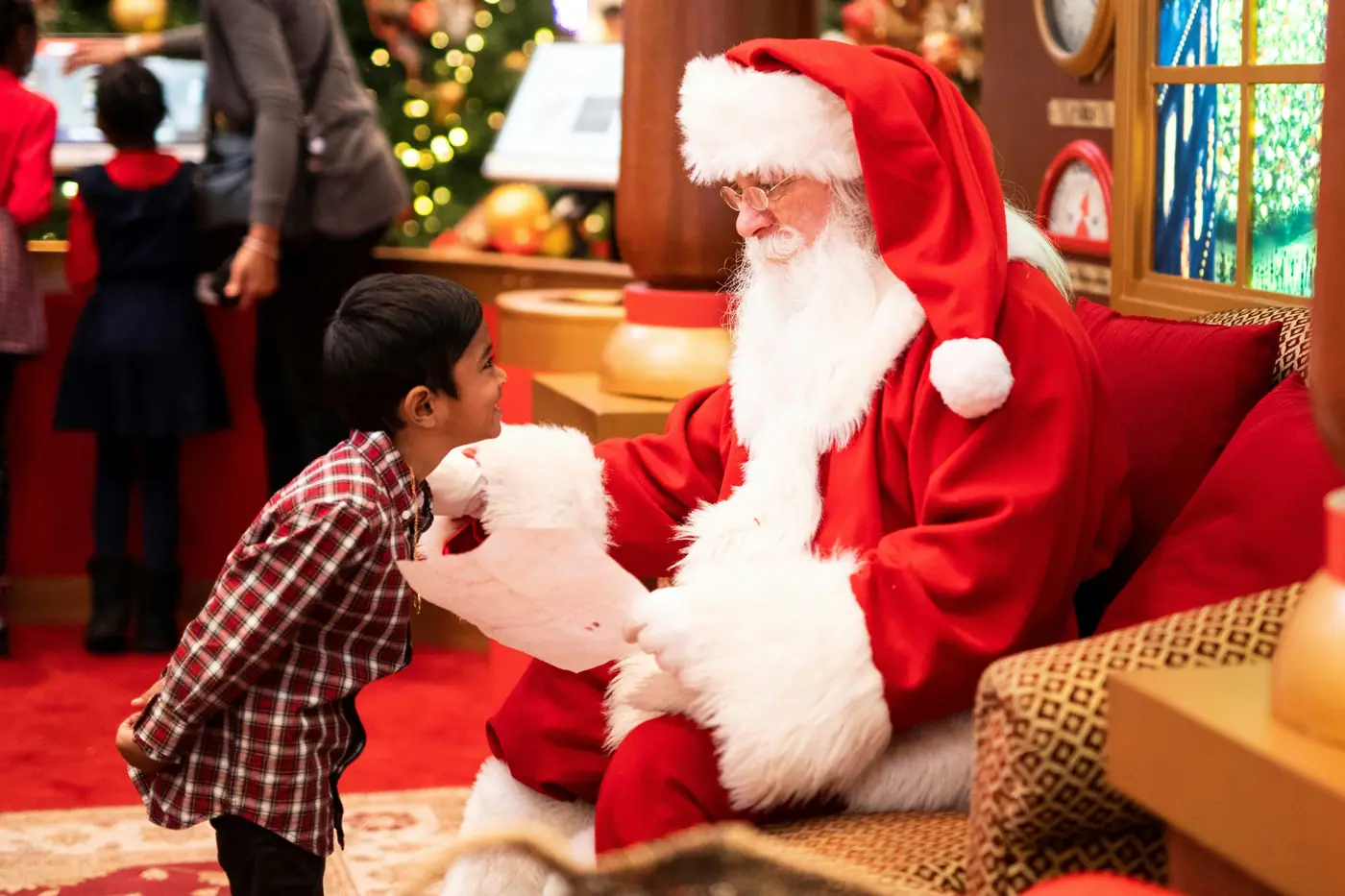 A small boy looking up at Santa Claus as he reads a letter, a Christmas tree glowing behind them
