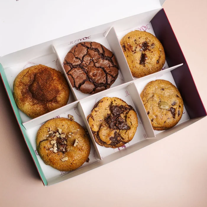 A box of six thick cookies, some topped with chocolate and nuts