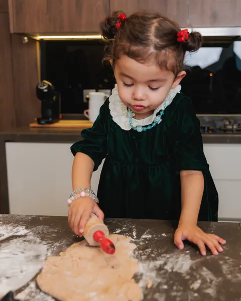 A small girl in a green velvet dress rolling dough with a red rolling pin