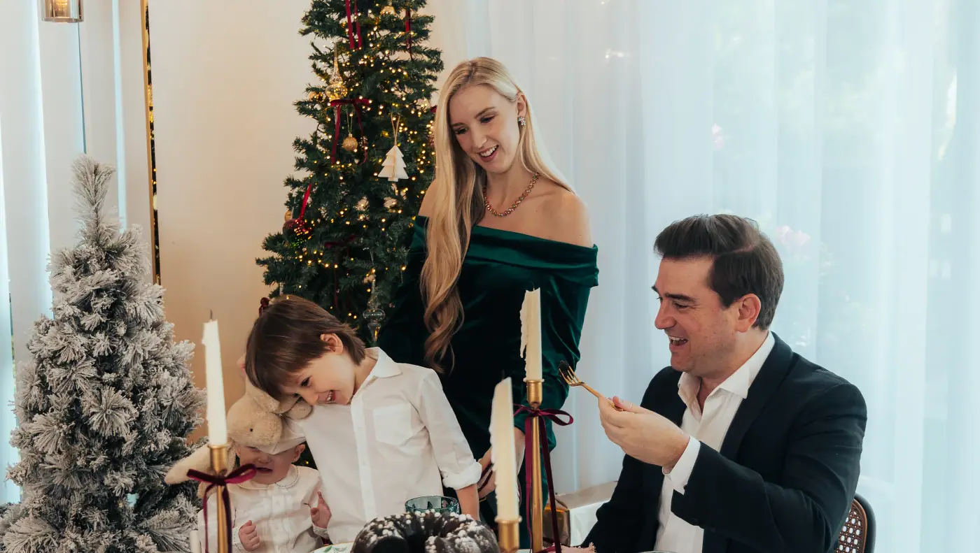 A family of four laughing around a Christmas table, two small children between their parents, a lit tree behind them
