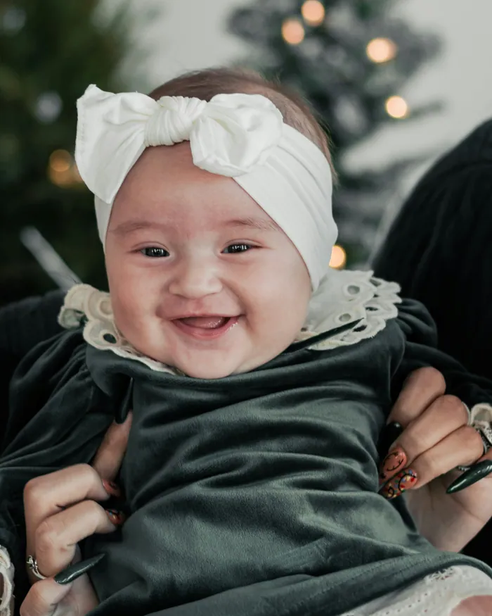 A smiling baby in a dark green velvet dress with a cream lace collar, held up in front of a lit Christmas tree