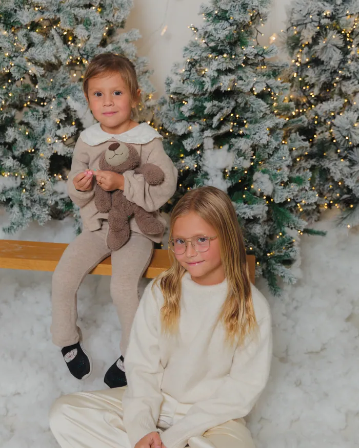 Two girls in cream sitting in the snow with a teddy bear between the flocked trees