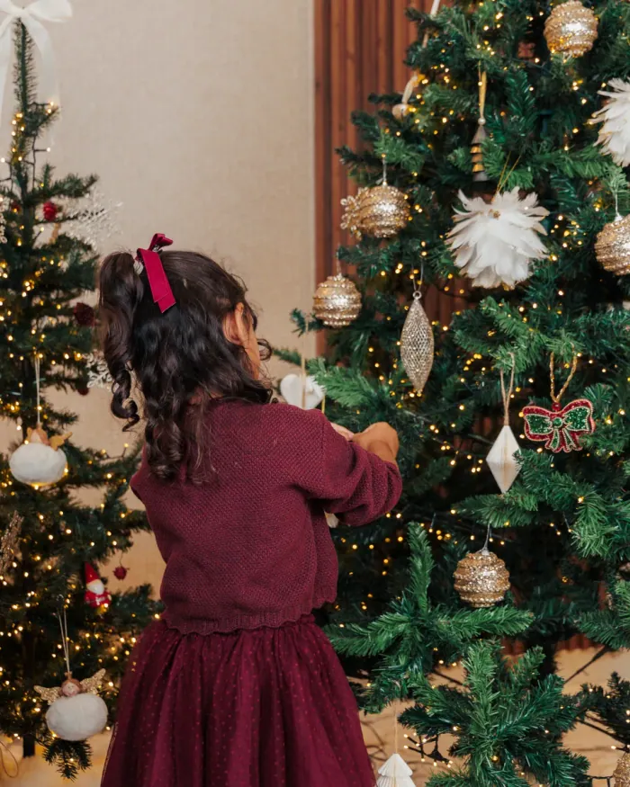 A small girl in a burgundy dress reaching up to hang an ornament on the tree