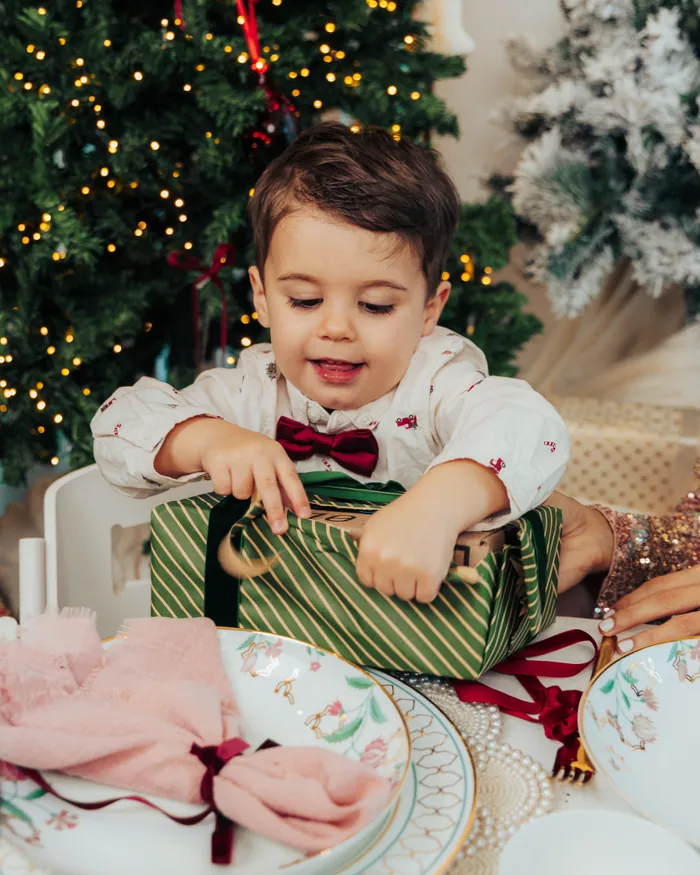 A small boy in a burgundy bow tie pulling the ribbon off a green striped present at the table