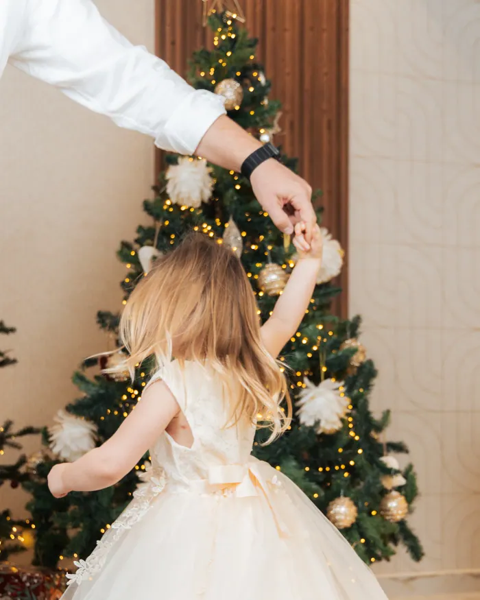 A small girl in a cream tulle dress spinning under her father&rsquo;s hand in front of a lit tree