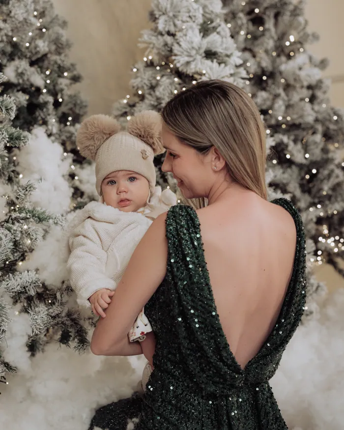 A mother in a deep green sequin dress turning to look at the baby she is holding, between two snowy trees