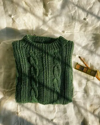 A deep green cable-knit jumper laid flat on cream linen beside a brass compact