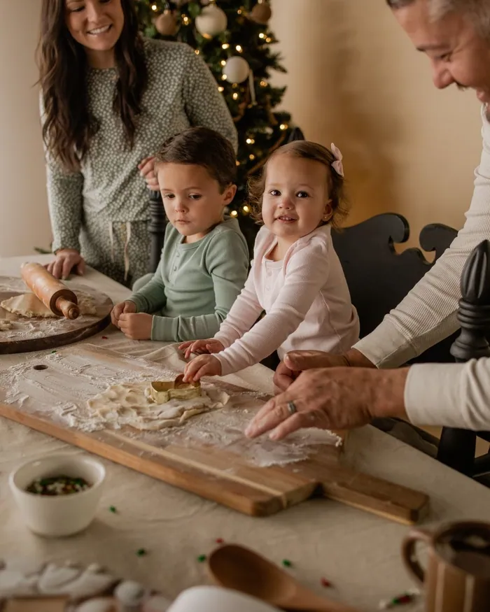 A family in soft green, pink and grey knits rolling out dough at a floury table by the tree