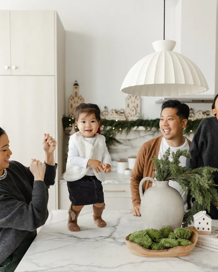 A toddler in a cream fleece and denim skirt dancing on a marble counter as her family laughs