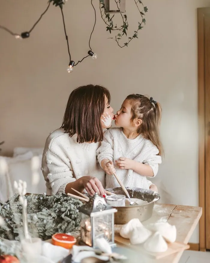 A mother and daughter in white knits sharing a kiss over a mixing bowl