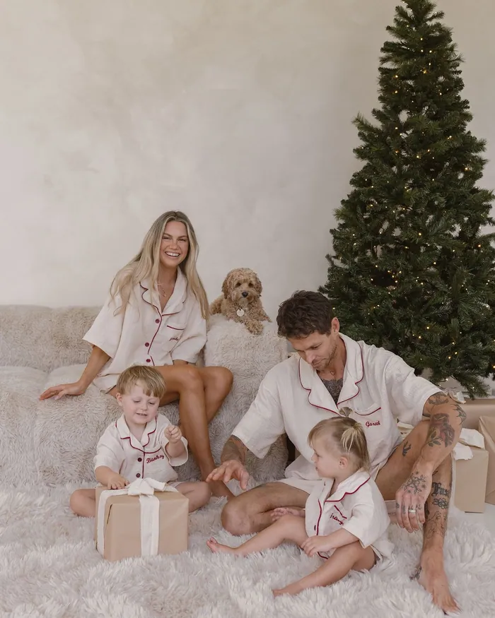 A family in white cotton pyjamas with fine piping on a fluffy rug beside the tree, a small dog behind them