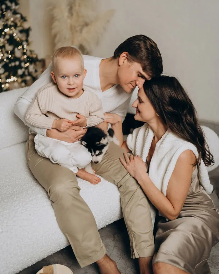 A family in soft knits, cream trousers and satin on a white sofa, the toddler holding a puppy