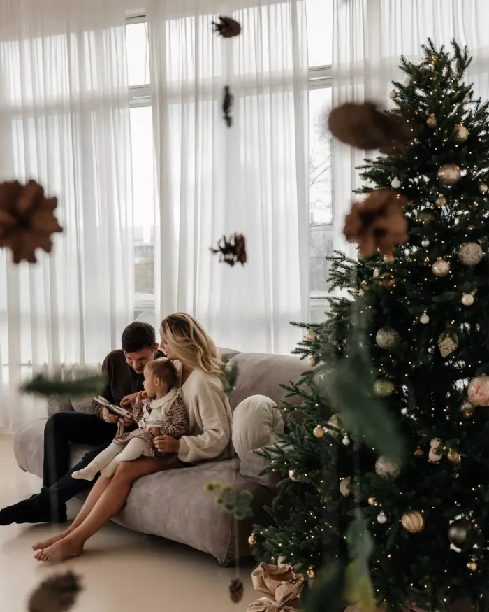 A family in soft knits curled up together on a grey sofa by a tall window, the tree beside them
