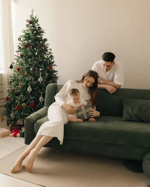 A mother in a cream knitted dress with her baby on a green sofa, the father leaning over the back, the tree beside them