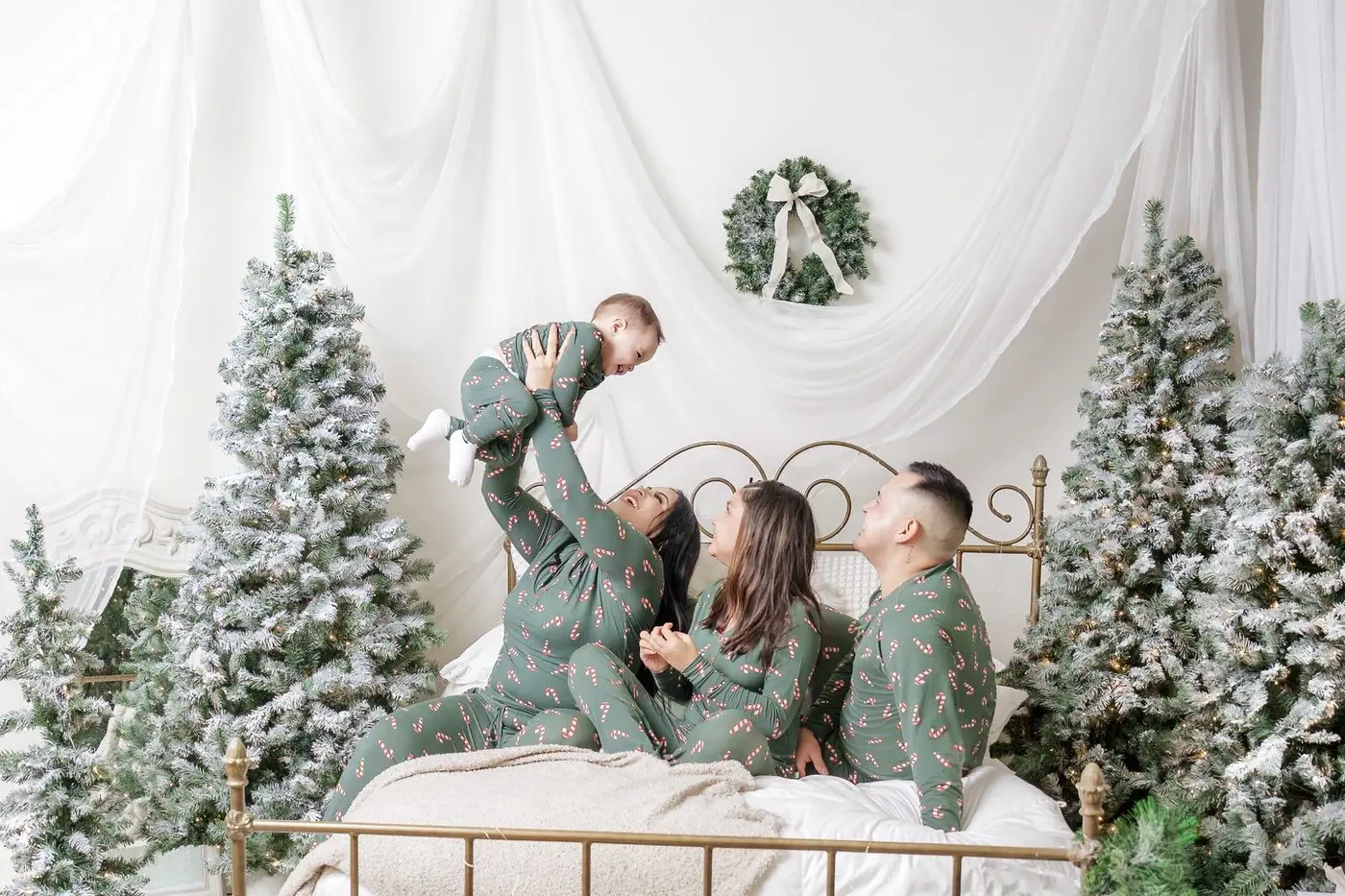 A family in matching green pyjamas on a brass bed between frosted trees, the father lifting the baby into the air