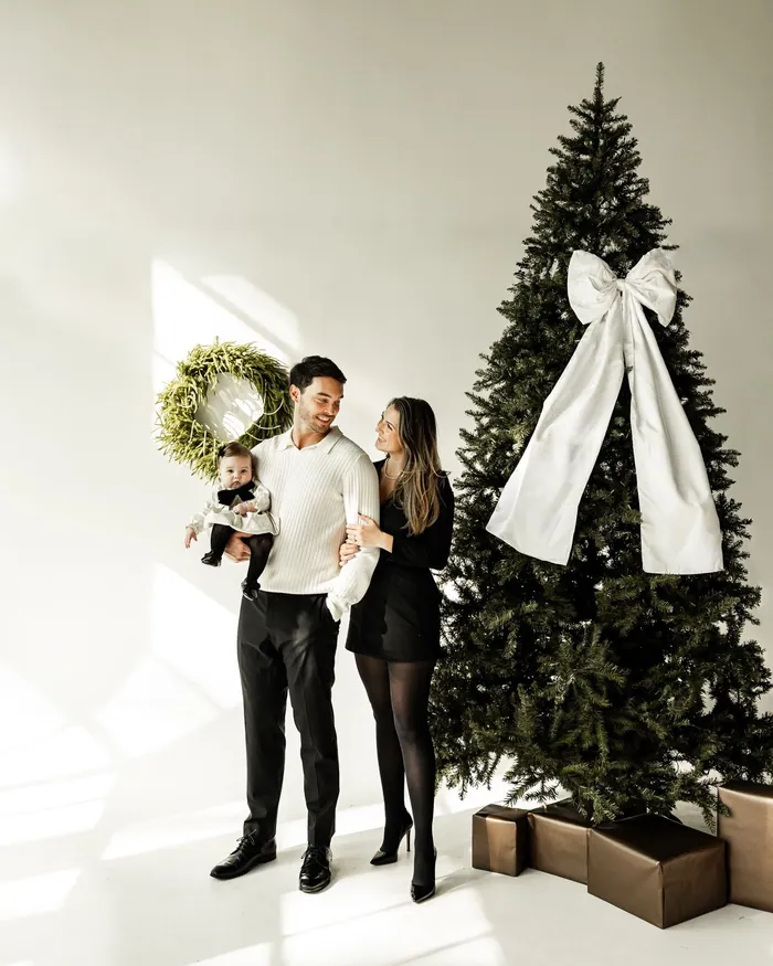 A father in a cream jumper and black trousers and a mother in a black dress holding their baby beside a tree tied with a white bow