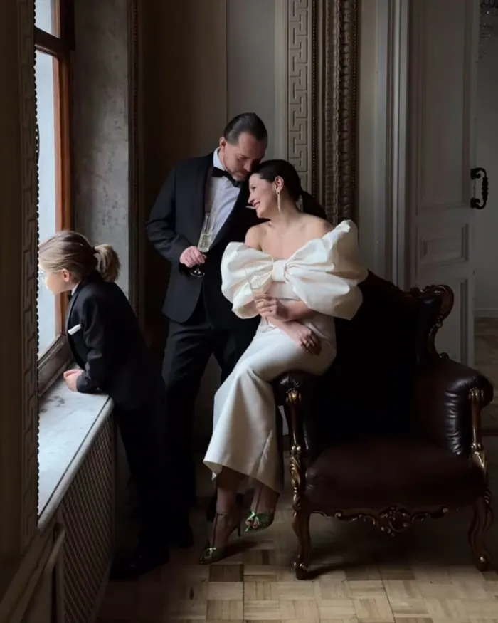 A couple in black tie and an ivory dress with a large bow, their child in a black suit looking out of the window