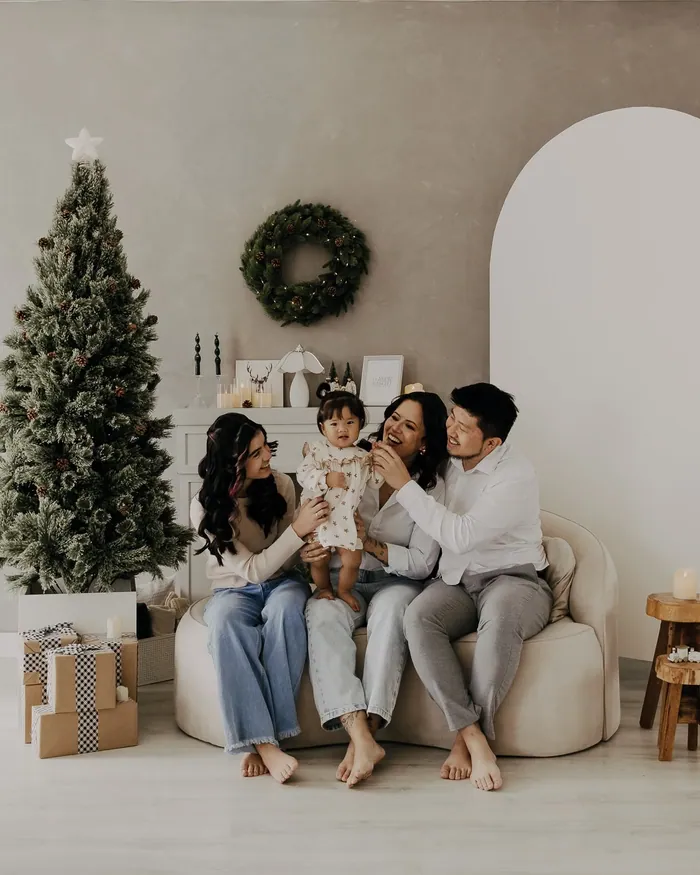 A family in light knits, white shirts and denim on a cream sofa beside a frosted tree