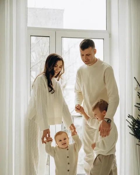 A mother and father in cream knits walking their baby across the floor by a bright window, an older boy beside them