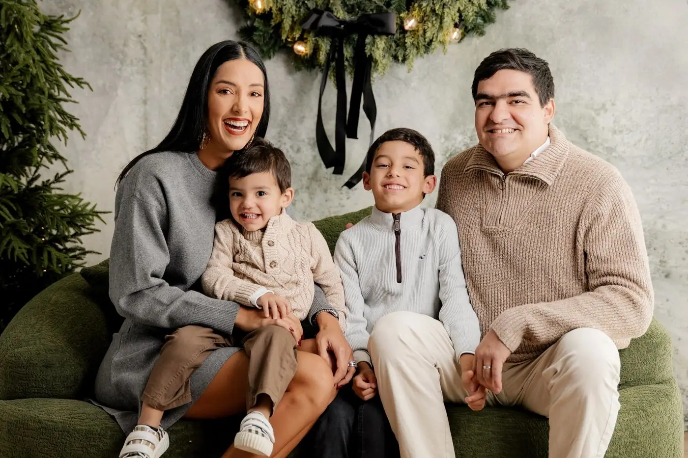 A family in grey, oatmeal and cream knits on a green sofa beneath a large wreath