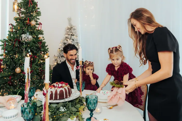 A family gathered around a laid Christmas table with a bundt cake and candles, two decorated trees behind them