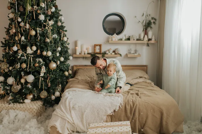 A father holding his baby up on a bed beside a tall Christmas tree hung with gold and white baubles