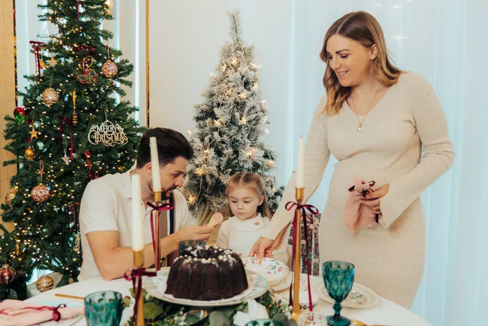 A family around a laid Christmas table with a chocolate bundt cake, the father leaning in to help his small daughter