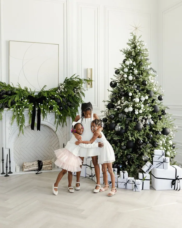 A fresh fir garland tied with a black velvet bow across a white fireplace, three small girls hugging beside the tree
