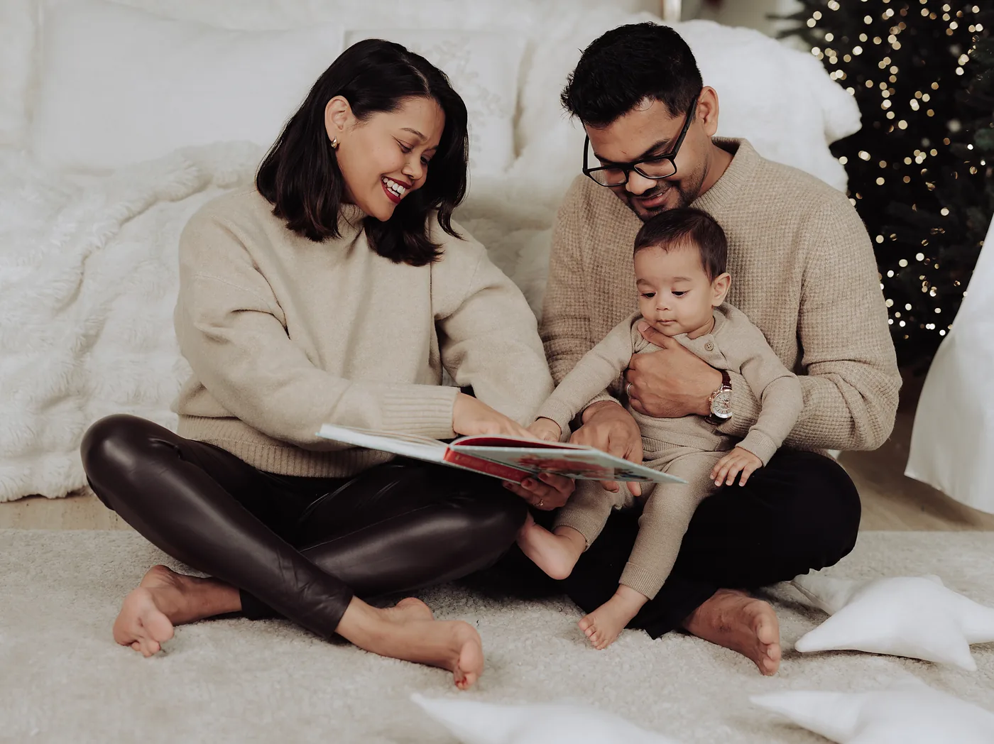 Two parents sitting on the floor reading a picture book to their baby, a lit tree behind them