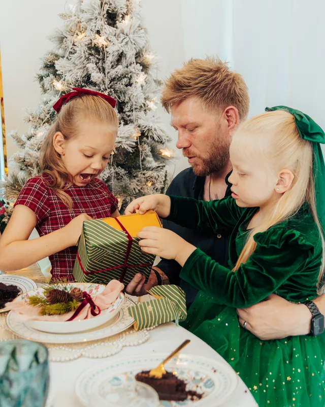 A father and his two daughters opening a wrapped gift at the table, the tree behind them
