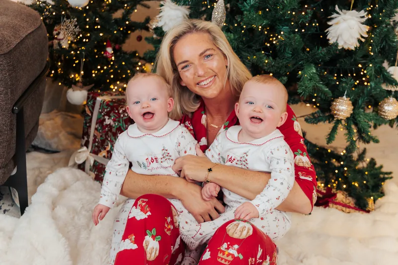 A mother sitting by the tree with her twin babies in Christmas pyjamas, one on each side