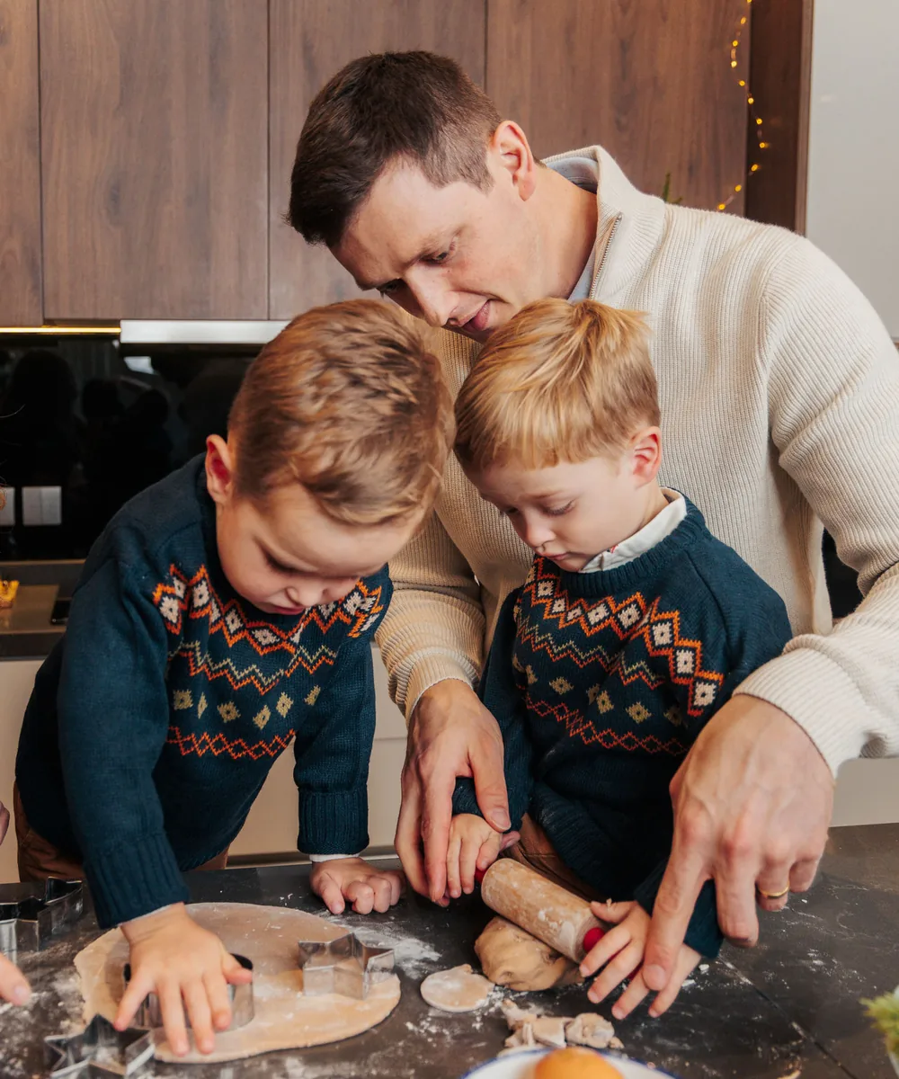 A father and two boys in fair isle jumpers cutting gingerbread shapes on a floured counter