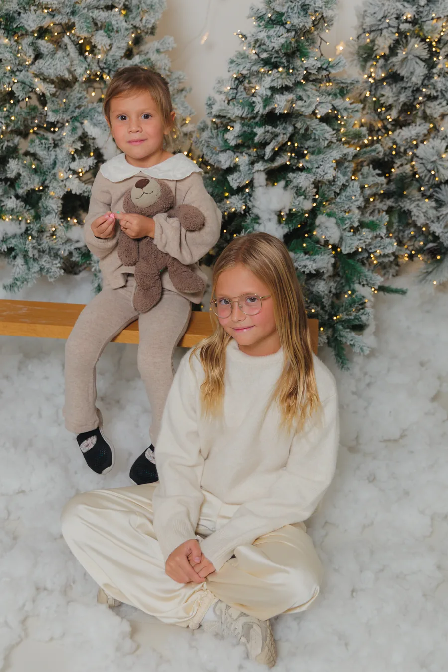 Two girls in cream sitting in the snow with a teddy bear between the flocked trees