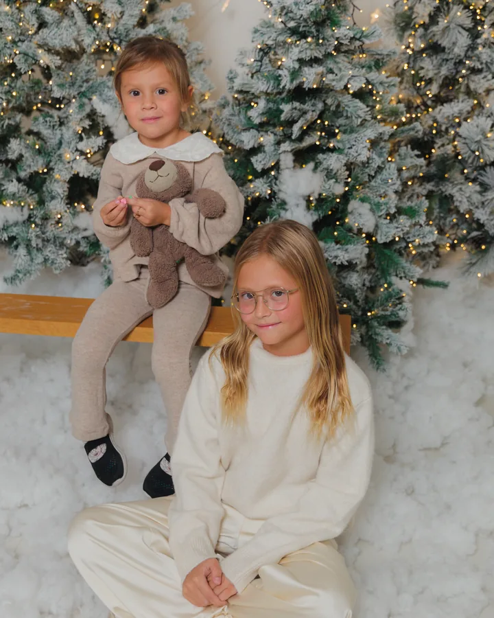 Two girls in cream sitting in the snow with a teddy bear between the flocked trees