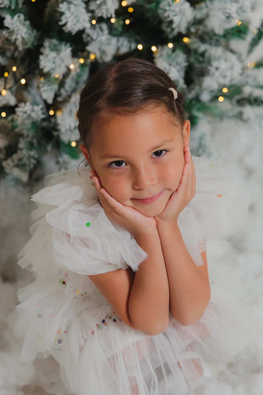 A girl in a white tulle dress with her chin in her hands, snow-covered branches behind her