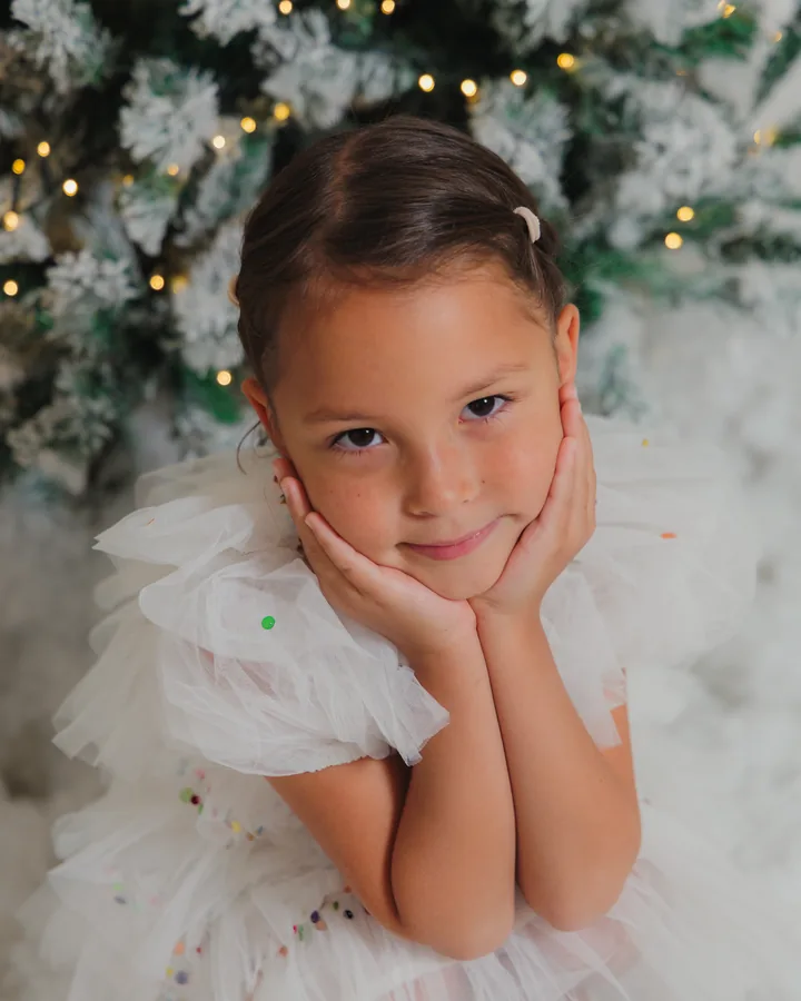 A girl in a white tulle dress with her chin in her hands, snow-covered branches behind her