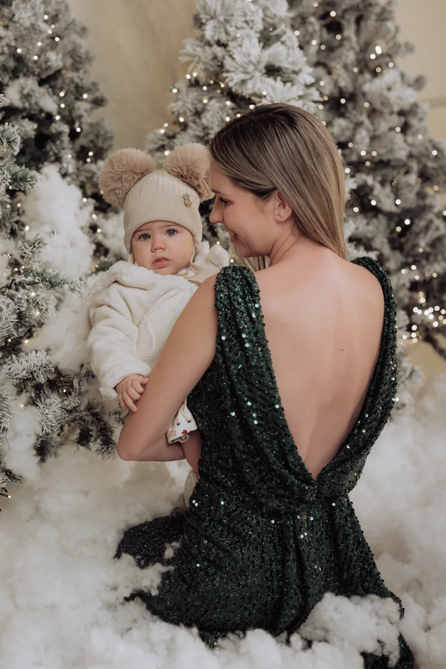 A mother in a green sequin dress holding her baby in a white bobble hat, seen from behind in the snow