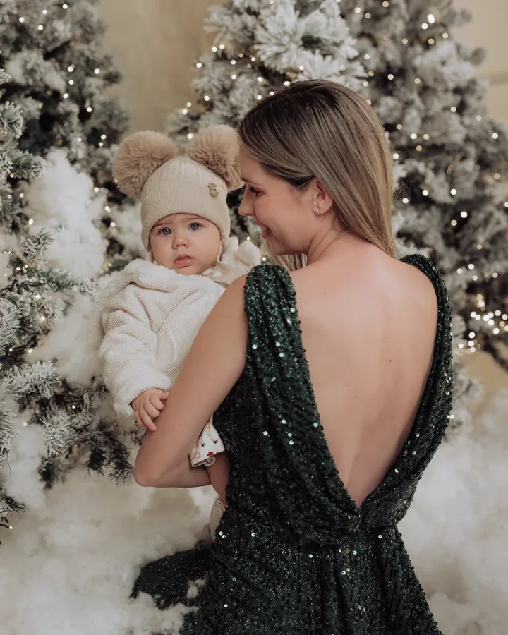 A mother in a green sequin dress holding her baby in a white bobble hat, seen from behind in the snow