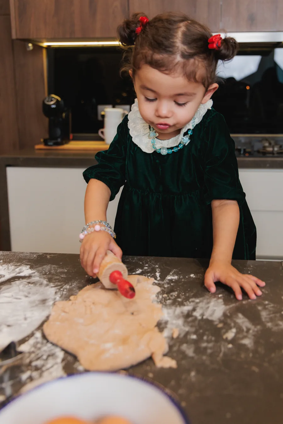 A small girl in a green velvet dress rolling dough with a red rolling pin