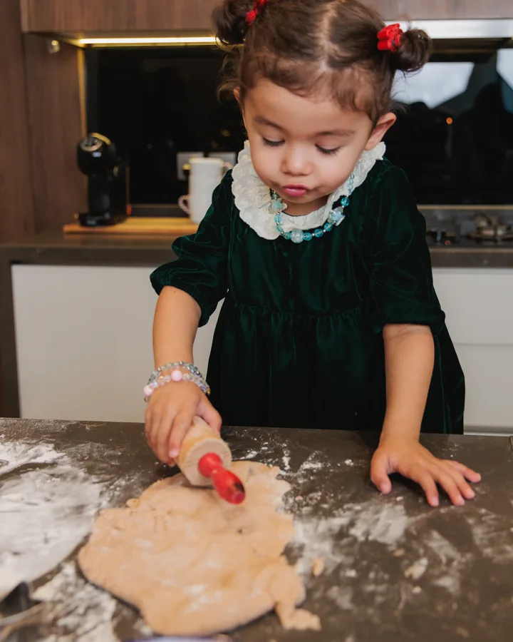 A small girl in a green velvet dress rolling dough with a red rolling pin